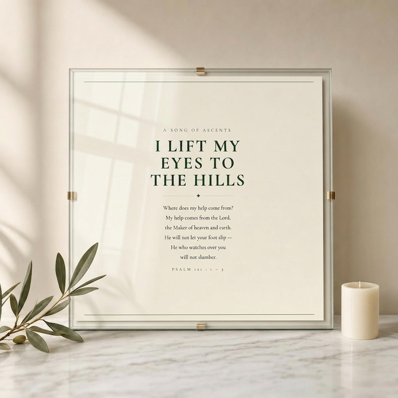 Psalm 121 I Lift My Eyes to the Hills scripture print in floating glass frame with brass corner clips on white marble surface with fresh olive branch and cream pillar candle in warm light, dark forest green text on warm cream background, premium christian wall art psalm 121 housewarming baptism gift