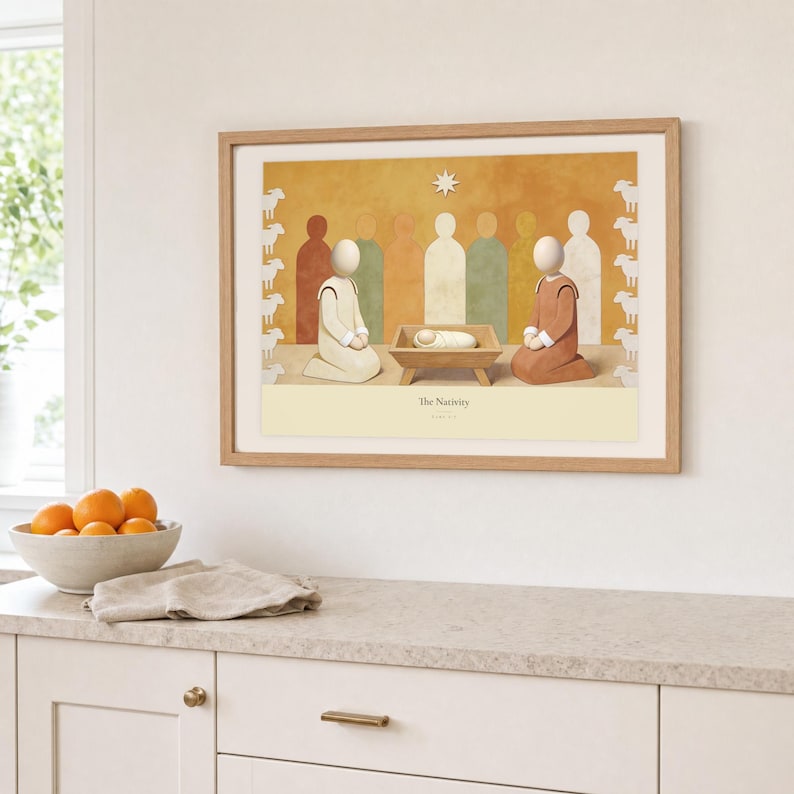 Kitchen mockup of The Nativity framed in light oak above a pale stone counter, white cabinetry, a linen cloth, and a bowl of oranges. The artwork shows the wrapped child in a low manger between two kneeling figures, beneath a single star. Bright window light and the warm cream mat demonstrate how the Christian print works in a clean, welcoming kitchen interior.