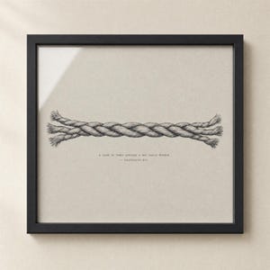 Hero close-up of the Cord of Three Strands wall art print in a matte black frame on a cream plaster wall, glass reflection visible. Hand-lettered Ecclesiastes 4:12 caption on warm grey paper. High-quality digital download for Christian wedding, marriage, or anniversary gift.