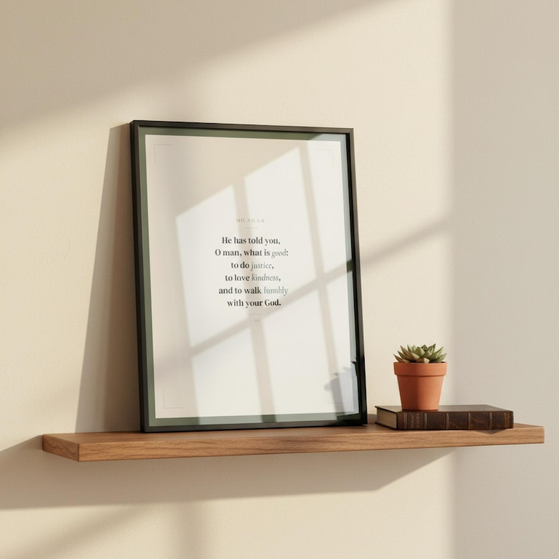 Micah 6:8 scripture wall art print in thin black glass frame leaning on natural wood floating shelf with small succulent plant and leather bound book beside it. Warm natural window light from left side. He has told you O man what is good to do justice to love kindness and to walk humbly with your God ESV. Sage green Christian Father Day gift digital download printable for home office study bedroom living room