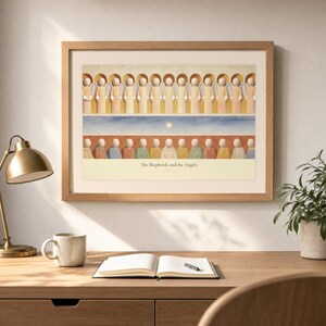 Desk mockup of The Shepherds and the Angels in a light oak frame above a wooden workspace with a brass lamp, ceramic mug, open notebook, pen, and leafy plant. The artwork shows a row of angels above shepherds and their sheep, with a star set between the two groups. Angled sunlight, neutral styling, and cream matting place the Bible story print in a quiet study or home office setting.