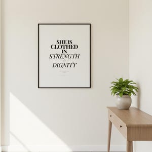 Proverbs 31 25 Wall Art She is Clothed in Strength and Dignity Scripture Print, — "She is clothed with strength and dignity, and she laughs without fear of the future", in elegant Mother's Day design with soft pink watercolor botanical flowers and graceful serif typography, photographed in beautiful close-up detail showing the premium glass frame reflection