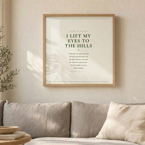 Psalm 121 I Lift My Eyes to the Hills scripture wall art print displayed above grey linen sofa in bright airy living room with olive branch in vase and palm leaf shadow, light oak wood frame, natural window light, christian minimalist home decor psalm 121 print baptism gift housewarming gift new home