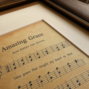 Closeup detail shot of Amazing Grace vintage hymn print on matte fine-art paper, showing the aged sepia hymnal paper texture, the serif AMAZING GRACE headline, the John Newton 1779 credit and the music notation in warm sepia tone. Premium vintage Christian sheet music print, instant digital download from TheFramedVerse on Etsy.