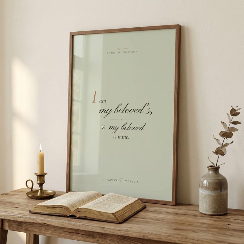 Song of Solomon 6:3 'I Am My Beloved's' coquette sage Christian wedding scripture print framed in pale oak, leaning on a cream console next to an open vintage Bible, brass candlestick with lit beeswax candle, ceramic vase with white peonies and dried wheat, romantic editorial newlywed Christian-faith bridal decor, TheFramedVerse digital download