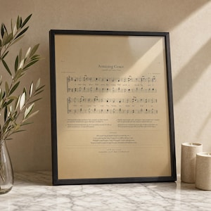 Amazing Grace vintage hymnal page printable in a slim warm-walnut frame, leaning on white Carrara marble next to a clear glass bud vase with a single fresh olive branch and an open vintage leather hymnal showing music notation pages. Premium Pinterest-worthy Christian sheet music print, instant digital download from TheFramedVerse on Etsy.