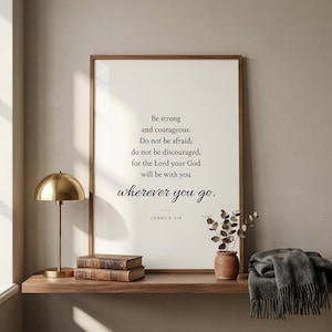 Joshua 1:9 'Be Strong and Courageous' Christian wall art in warm walnut frame leaning on walnut shelf beside leather Bible stack, brass desk lamp, and eucalyptus stems. Editorial masculine Christian home interior. Father's Day gift for Christian dads and leaders, confirmation and graduation print, instant digital download.