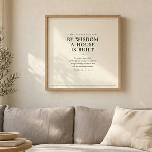Proverbs 24:3 By Wisdom A House Is Built scripture wall art print displayed above grey linen sofa in airy living room with olive branch, light oak frame, natural window light and palm leaf shadows, christian minimalist home decor, house blessing print housewarming gift scripture wall art entryway new home