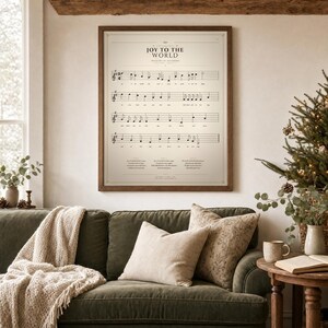 Joy to the World vintage hymn print featuring Psalm 98:4 in an oak frame above a sage-green linen sofa with a knit throw and cream pillows, a pine sprig and soft warm window light, cozy farmhouse living room. Single holiday Christian sheet music wall art download from TheFramedVerse Etsy, the carol on a warm sepia hymnal page in cream and antique black.