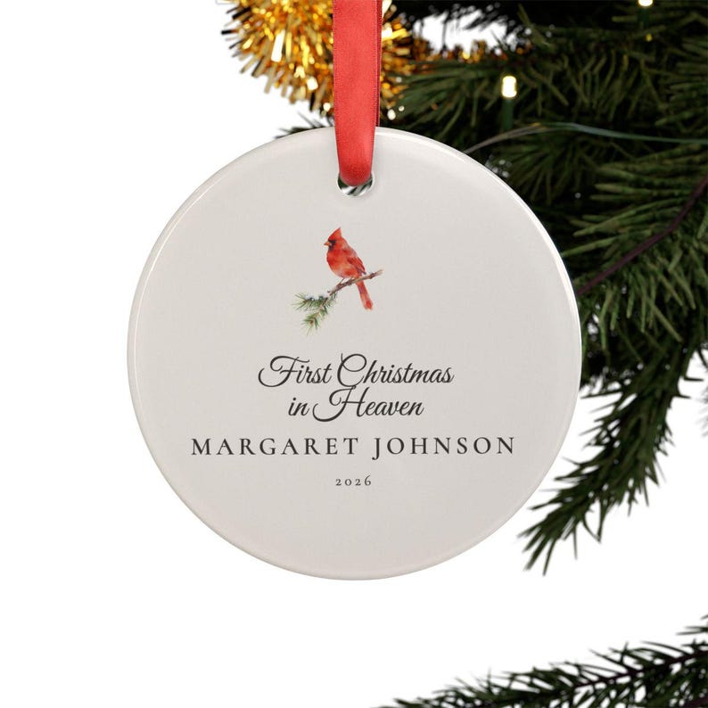 Christmas tree mockup showing a round personalized First Christmas in Heaven ornament hanging from a ribbon among snowy evergreen branches and warm lights. The clear acrylic keepsake features a red cardinal on a snow covered pine branch, the name Margaret Johnson and the year 2026, creating a heartfelt memorial ornament for remembering a loved one during the holidays.