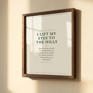 Close-up detail of Psalm 121 I Lift My Eyes to the Hills scripture print in warm walnut float frame with glass and natural golden window light casting soft shadows, dark forest green Cormorant Garamond serif typography on warm cream paper, christian wall art close-up lift my eyes psalm 121 print housewarming baptism gift