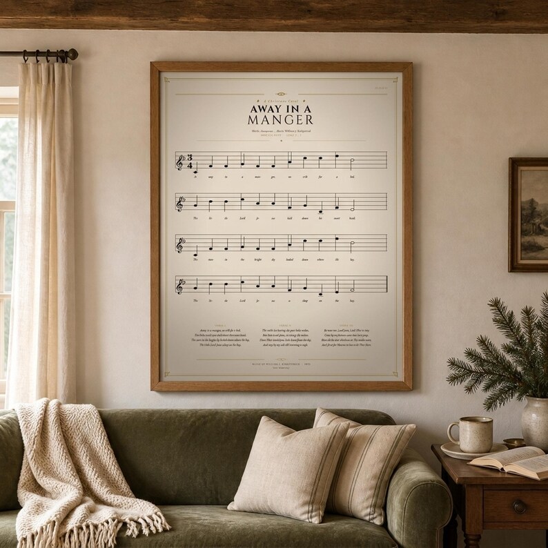 Away in a Manger vintage hymn print featuring Luke 2:7 in an oak frame above a sage-green linen sofa with a knit throw and cream pillows, a pine sprig and soft warm window light, cozy farmhouse living room. Single holiday Christian sheet music wall art download from TheFramedVerse Etsy, the carol on a warm sepia hymnal page in cream and antique black.