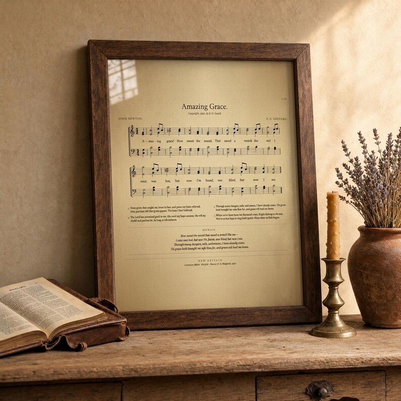 Amazing Grace vintage hymn print framed in rustic walnut wood, leaning on a reclaimed-oak console next to an open vintage leather hymnal Bible, antique brass candlestick with lit beeswax candle, and terracotta vase with dried wheat and lavender. Reverent rustic Christian-faith home decor, instant digital download from TheFramedVerse on Etsy.