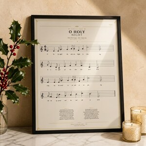 O Holy Night vintage sheet music print in a thin black wood frame on a white Carrara marble surface, a single holly branch with berries casting a soft shadow, two cream unlit votive candles, warm beige wall backdrop, bright winter light with glass reflection. Editorial still life Christmas carol hymn print digital download TheFramedVerse.