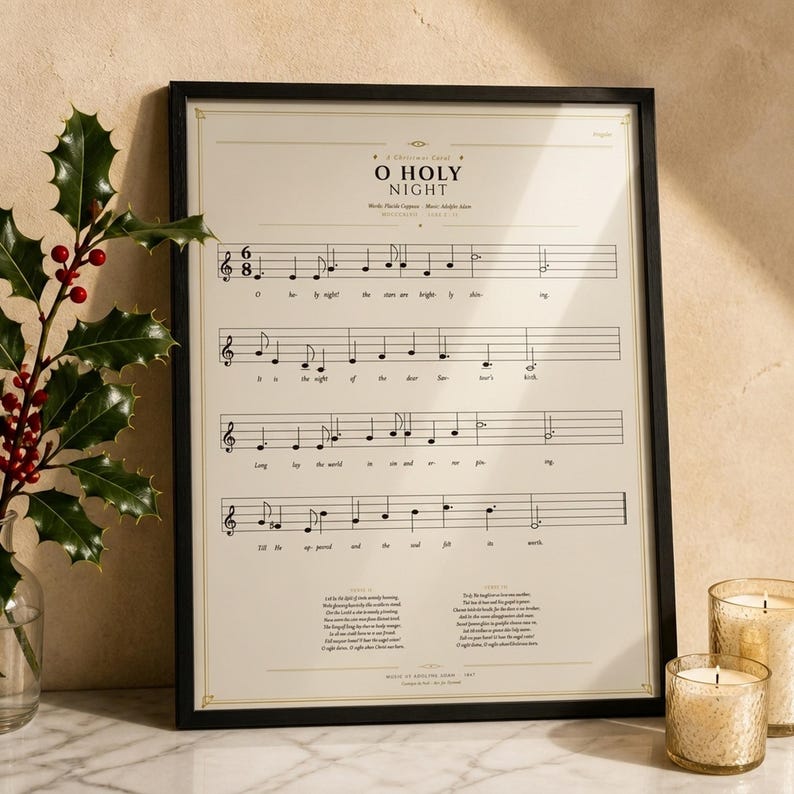 O Holy Night vintage sheet music print in a thin black wood frame on a white Carrara marble surface, a single holly branch with berries casting a soft shadow, two cream unlit votive candles, warm beige wall backdrop, bright winter light with glass reflection. Editorial still life Christmas carol hymn print digital download TheFramedVerse.