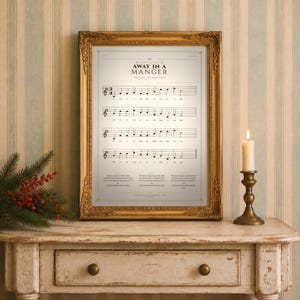 Away in a Manger vintage hymn print featuring Luke 2:7 framed in light oak resting on a wooden console with stacked books, pinecones and dried eucalyptus by a sunlit window, cozy farmhouse living room. Single Christmas sheet music wall art digital download from TheFramedVerse Etsy shop, aged sepia hymnal page style in cream and soft black tones.