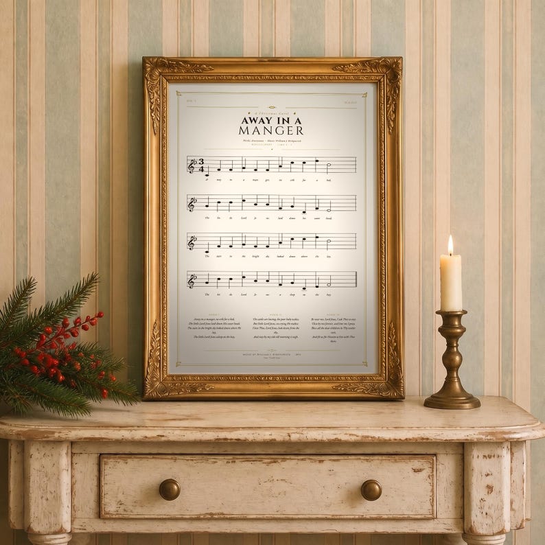 Away in a Manger vintage hymn print featuring Luke 2:7 framed in light oak resting on a wooden console with stacked books, pinecones and dried eucalyptus by a sunlit window, cozy farmhouse living room. Single Christmas sheet music wall art digital download from TheFramedVerse Etsy shop, aged sepia hymnal page style in cream and soft black tones.
