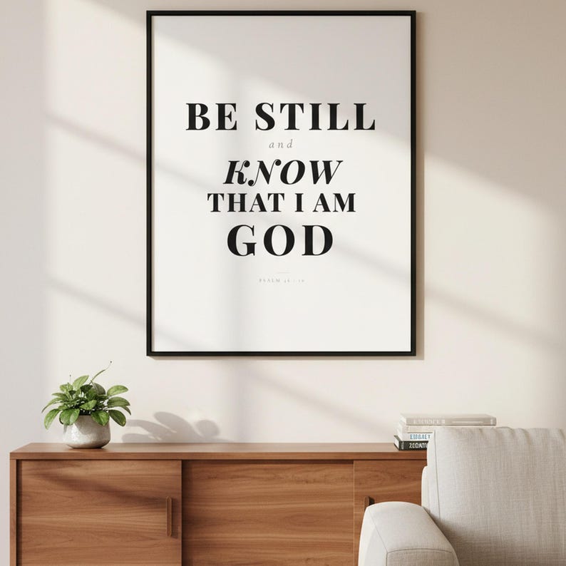 Psalm 46 10 Wall Art Be Still and Know That I Am God Scripture Print Christian, — "Be still and know that I am God. I will be exalted among the nations", in contemporary bold serif typography in black on clean white background, displayed in a beautifully styled modern living room above a warm wooden sideboard with soft natural light creating gentle shadows on the frame