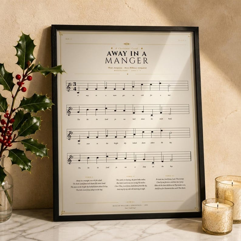 Away in a Manger vintage sheet music print in a thin black wood frame on a white Carrara marble surface, a single holly branch with berries casting a soft shadow, two cream unlit votive candles, warm beige wall backdrop, bright winter light with glass reflection. Editorial still life Christmas hymn print digital download TheFramedVerse.