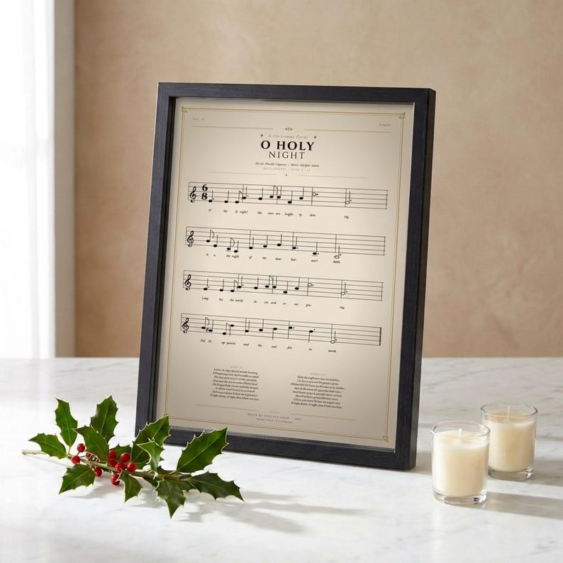 O Holy Night vintage sheet music print in thin black wood frame on white Carrara marble surface, single holly branch with berries casting soft shadow, two cream unlit votive candles, warm beige wall backdrop, bright winter light with glass reflection, editorial still life christmas carol hymn print digital download TheFramedVerse