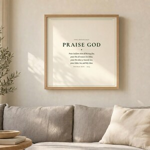 Doxology Praise God From Whom All Blessings Flow hymn lyric wall art print above grey linen sofa in bright airy living room with olive branch, light oak wood frame, natural window light and palm shadows, christian minimalist home decor, doxology print song lyric art baptism confirmation wedding gift