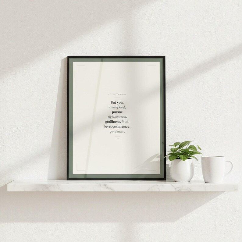 1 Timothy 6:11 Man of God scripture wall art print in black glass frame on white marble shelf with green potted plant and white coffee mug. Window light creating diagonal shadow. But you man of God pursue righteousness godliness faith love endurance gentleness ESV. Sage green Christian Father Day gift for dad husband pastor digital download printable wall art for home office bedroom living room