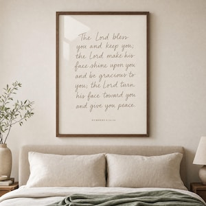 Hand-lettered Numbers 6:24-26 priestly blessing print in a honey-walnut frame hanging above an oat-linen upholstered headboard with two cream linen pillows, soft morning window light reflecting on the glass. Modern Christian wedding scripture bedroom wall art, instant digital download for marriage and anniversary gifts.
