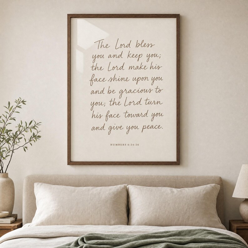 Hand-lettered Numbers 6:24-26 priestly blessing print in a honey-walnut frame hanging above an oat-linen upholstered headboard with two cream linen pillows, soft morning window light reflecting on the glass. Modern Christian wedding scripture bedroom wall art, instant digital download for marriage and anniversary gifts.