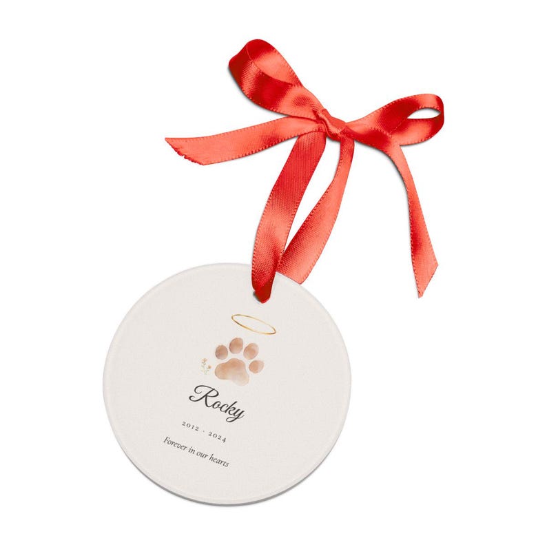 Close hero view of a personalized pet memorial ornament held gently in one hand. The round clear acrylic keepsake shows a watercolor Dog motif, the name Rocky and the years 2012 to 2024, with a ribbon threaded through the top. Natural light reveals the smooth edge, vivid double sided print and thoughtful scale of the finished dog memorial gift.