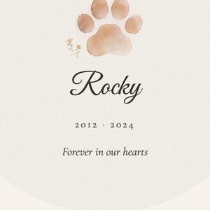 Detailed close up of the custom pet name and memorial years printed on the acrylic ornament. Crisp lettering shows Rocky and 2012 to 2024 beneath the soft watercolor Paw with Halo artwork, while the rounded edge and glossy surface catch gentle natural light. The tight crop highlights the readable personalization and careful finish of the pet remembrance keepsake.
