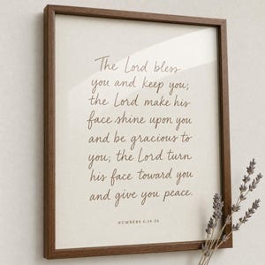 Premium close-up of the Numbers 6:24-26 hand-lettered priestly blessing in a warm oak frame, slightly angled with a soft sprig of dried wildflowers in the foreground bokeh and clear glass reflection. Aaronic blessing 'The Lord bless you' Christian wedding bible verse wall art, instant digital download.