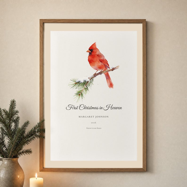 Lifestyle holiday room mockup showing a personalized First Christmas in Heaven print in a warm wood frame above a softly decorated console. A red cardinal rests among snow covered pine branches above Margaret Johnson and 2026. Gentle lights, evergreen accents and neutral decor show how this memorial Christmas gift brings a quiet remembrance into a family home.