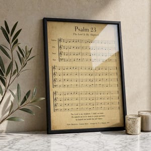 Psalm 23 psalm 23 print in thin black frame with visible glass front, leaning on white Carrara marble surface, single olive branch on left casting soft shadow, two small votive candles on right, warm beige textured wall backdrop, bright morning light with clean glass reflection, TheFramedVerse Etsy psalms 23
