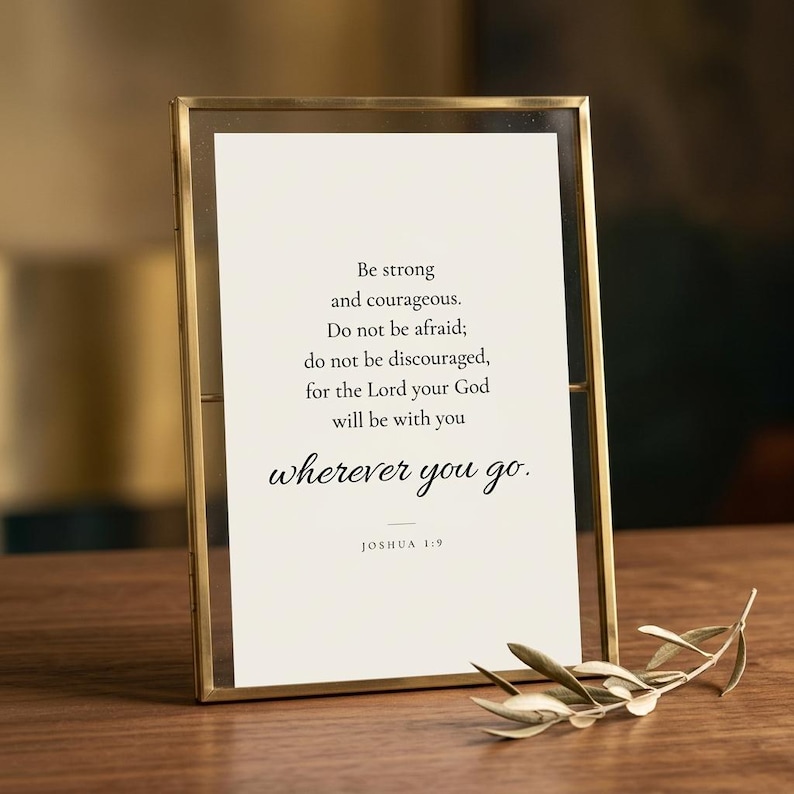 Joshua 1:9 'Be Strong and Courageous' scripture print in elegant brass-edged glass float frame with realistic glass surface reflections, beside dried olive sprig on warm walnut surface. Botanical editorial product photography, Father's Day Christian wall art gift, confirmation, baptism, graduation keepsake, instant download.