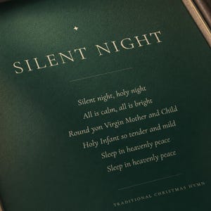 Angled close-up of the Silent Night Christmas print focusing on the cream serif SILENT NIGHT title and delicate gold star ornament against deep forest green. Part of the centered first hymn verse is visible below, highlighting the refined typography and subtle detail of this minimalist holiday print and timeless Christmas hymn wall art.