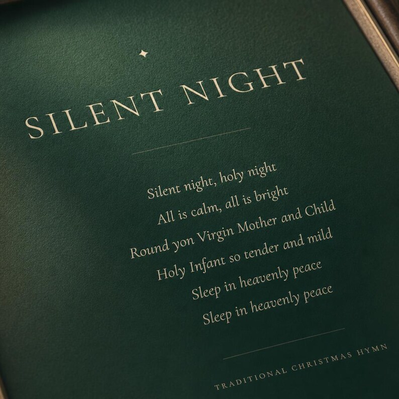 Angled close-up of the Silent Night Christmas print focusing on the cream serif SILENT NIGHT title and delicate gold star ornament against deep forest green. Part of the centered first hymn verse is visible below, highlighting the refined typography and subtle detail of this minimalist holiday print and timeless Christmas hymn wall art.