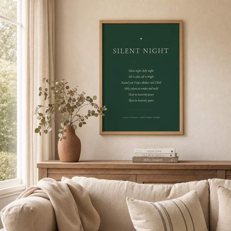 Framed Silent Night Christmas print displayed above a warm wood sideboard in a bright living room, styled with a vase of dried flowers. The deep forest green artwork features cream serif lettering, a gold star ornament, the hymn's first verse, and a Traditional Christmas Hymn footer, creating elegant Christian Christmas decor and minimalist holiday wall art.