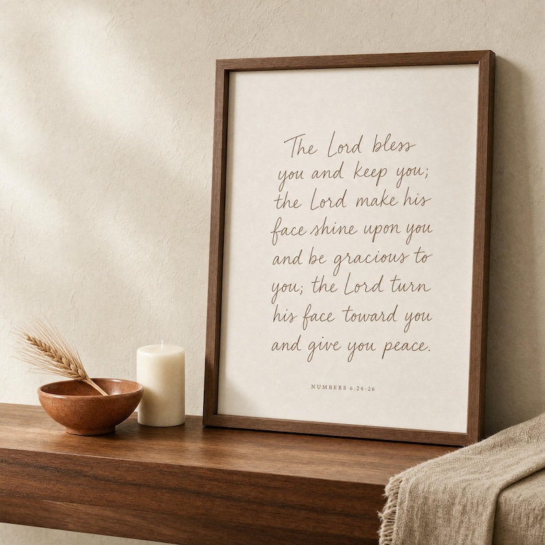 Numbers 6:24-26 Aaronic blessing print framed in warm walnut wood, leaning on a light olive-wood floating shelf next to a terracotta ceramic bud vase with dried wheat, two linen-bound books, and a brass bowl. Hand-lettered Christian wedding bible blessing wall art, premium digital download for housewarming and anniversary gifts.