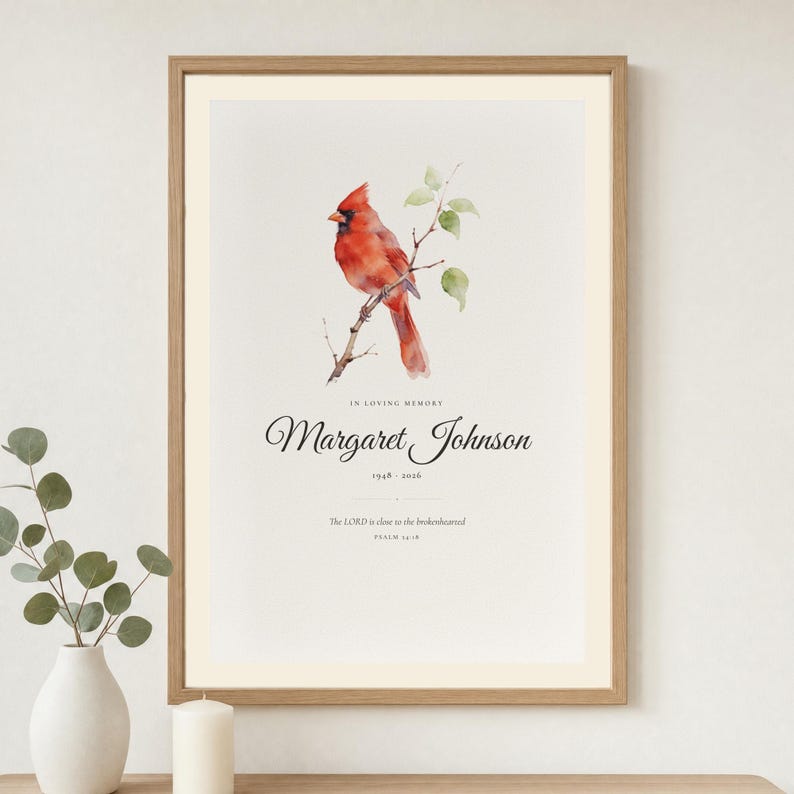 Lifestyle room mockup showing a personalized memorial print in a warm wood frame above a quiet neutral console. The artwork features a red cardinal, In Loving Memory, Margaret Johnson, 1948 to 2026, and Psalm 34:18, The Lord is close to the brokenhearted. Soft daylight and simple ceramic decor present a comforting sympathy gift for loss of a mother or father.