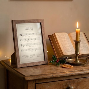 Away in a Manger vintage hymnal print in rustic walnut frame leaning on antique console, open vintage hymnal book beside, brass candlestick with beeswax candle, sprig of holly and dried orange slice, warm golden light, cottage Christmas faith home decor, TheFramedVerse digital download classic christmas hymn print