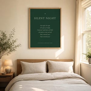 Framed Silent Night Christmas print hanging above a beige sofa with soft neutral cushions. The deep forest green artwork features cream serif lettering, a gold star ornament, the centered first verse, and a Traditional Christmas Hymn footer. This serene minimalist holiday print adds classic Christmas hymn wall art to a calm, welcoming living room.