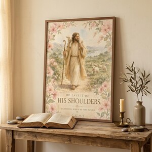 He Lays It On His Shoulders Luke 15:5 watercolor christian wall art leaning on a rustic console table beside an open vintage Bible, brass candlestick, and dried olive branch. Reverent Christian home decor styling — perfect for baptism gift, pastor appreciation, christian housewarming. Digital download.