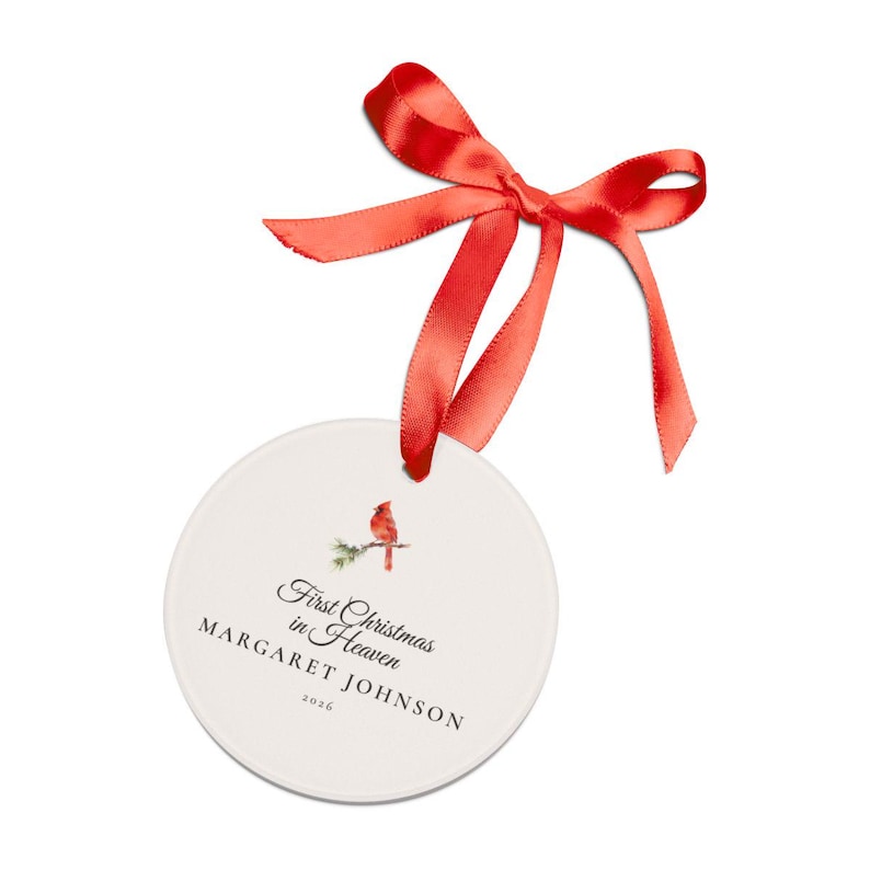 Neutral flatlay of a personalized First Christmas in Heaven ornament arranged with ribbon, evergreen sprigs and simple holiday details. The round acrylic front shows a watercolor cardinal on snowy pine above Margaret Johnson and 2026. The clean overhead view presents the ornament's size, circular shape and soft cream design as a remembrance gift for Christmas.