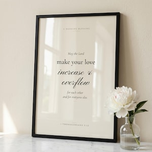 1 Thessalonians 3:12 'May the Lord Make Your Love Increase' Christian wedding scripture print in thin black wood frame on white Carrara marble, single olive branch casting soft shadow, two unlit votive candles, warm beige textured wall, bright morning light with clean glass reflection, editorial still life print download from TheFramedVerse