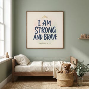 I Am Strong And Brave Joshua 1:9 print in modern boys nursery with sage green walls, wooden toddler bed, woven basket, and stuffed bear. Bold navy hand-lettered affirmation with tiny gold mountain icon. Part of Kids Christian wall art set of 6 — ideal baby boy baptism gift, boy nursery decor, christian gift.