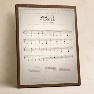 Away in a Manger vintage sheet music print in a solid walnut frame, frontal studio shot on a cream backdrop with soft top-left key light and gentle glass reflection. Classical Christmas hymn wall art digital download from TheFramedVerse — single hymnal page reproduction of Luke 2:7 in warm sepia, cream, and soft black tones.