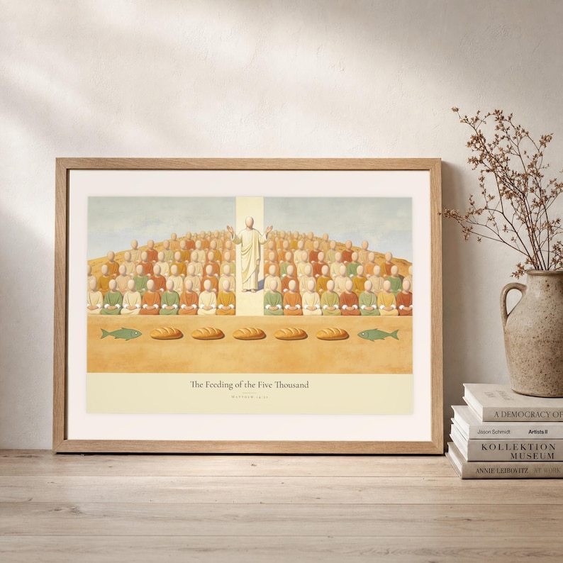 Leaning frame mockup of The Feeding of the Five Thousand on a rustic wood console beside stacked art books and a tall ceramic vessel with dried stems. The print shows Christ raised on the centre axis above a seated hillside crowd, with loaves and fish laid out along the foreground. The oak frame, broad cream mat, plaster wall, and soft side light create a relaxed display while keeping the title and Matthew 14:21 legible.