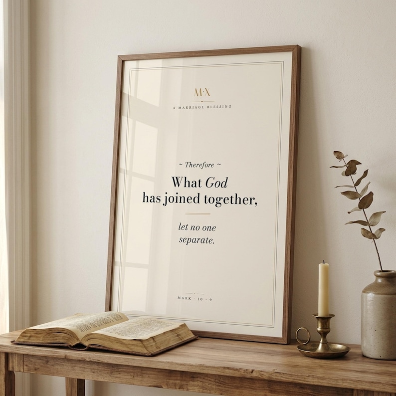 Mark 10:9 modern Christian wedding scripture print framed in rustic walnut wood, leaning on a rustic console next to an open vintage Bible, brass candlestick with lit beeswax candle, terracotta vase with dried wheat and white roses, cottage editorial newlywed Christian-faith home decor, TheFramedVerse digital download