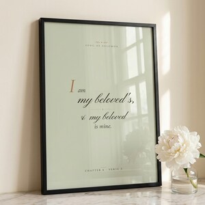 Song of Solomon 6:3 'I Am My Beloved's' coquette sage Christian wedding scripture print in thin black wood frame on white Carrara marble, single sage olive branch and white peony casting soft shadow, two unlit votive candles, warm beige textured wall, bright morning light with clean glass reflection, editorial bridal still life print download from TheFramedVerse