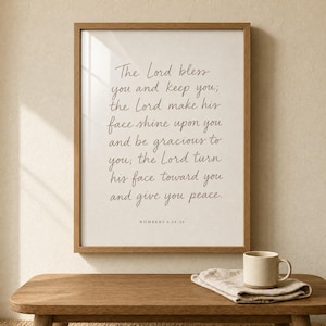 May include: A framed print with a light wood frame displays a script of text from Numbers 6:24-26. The text is in a handwritten style, in a dark brown colour, on a cream-coloured background. A beige mug and folded cloth sit on a wooden table.